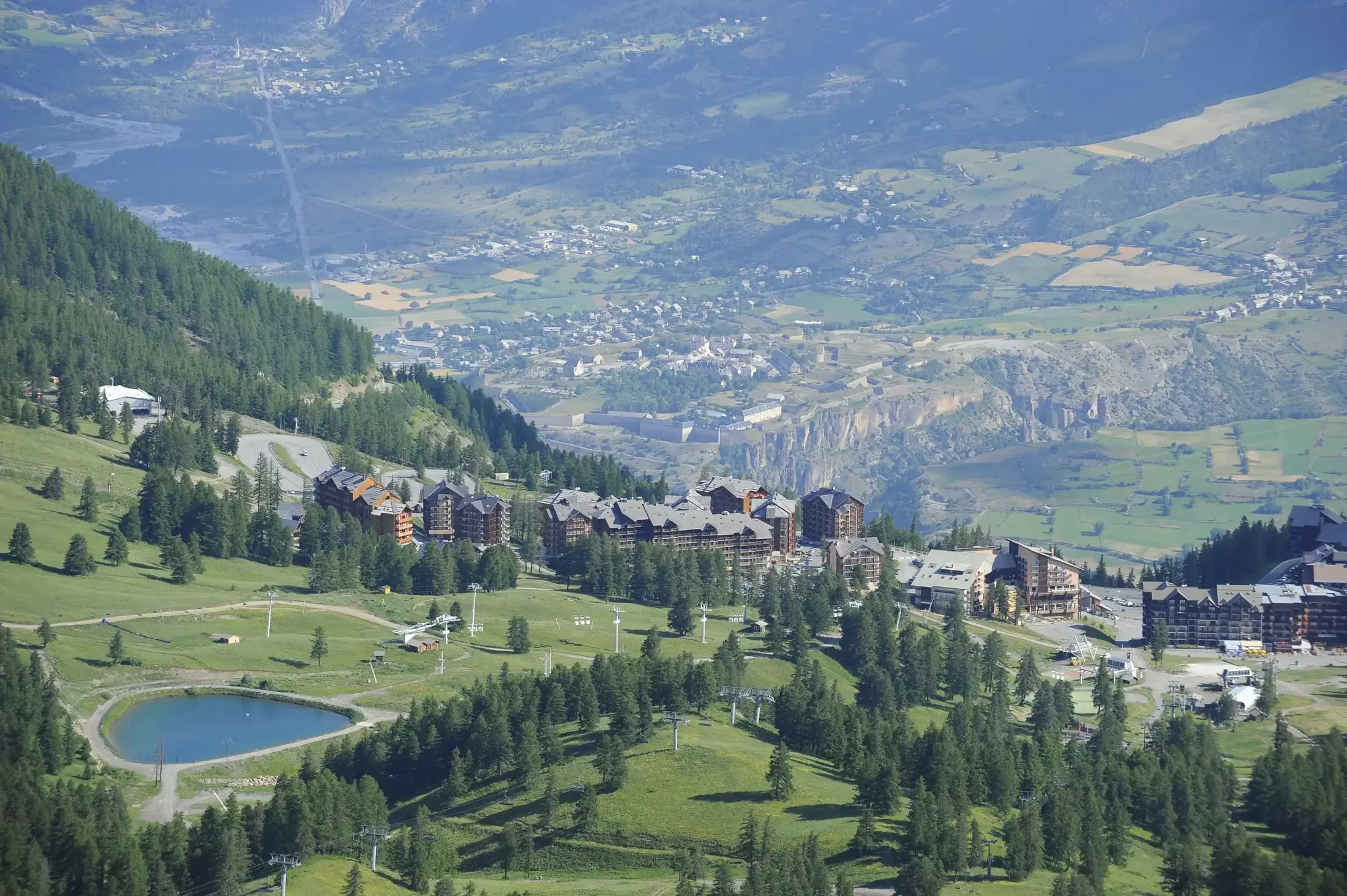 Ancelle en été : pistes redevenues prairies, plan d'eau, mélèzes et vallée du Champsaur en contrebas