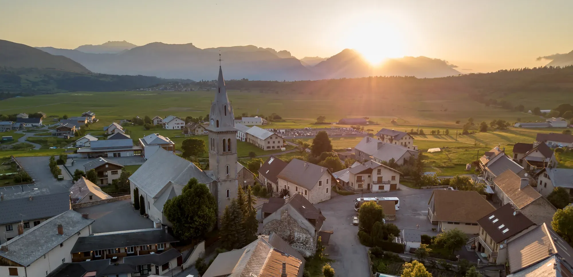 Le village d'Ancelle vu du ciel au coucher du soleil : le clocher, les toits et les prés du Champsaur, sommets du Dévoluy à l'horizon
