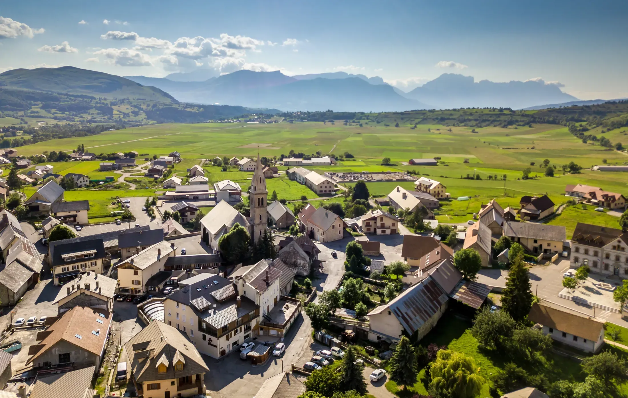 Le village d'Ancelle vu du ciel en été : toits d'ardoise, clocher et prairies du plateau du Champsaur, massifs du Dévoluy à l'horizon